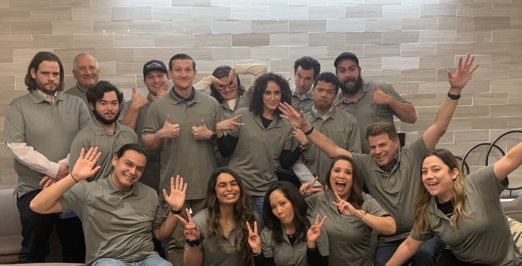 A cheerful group photo of Excellos team members wearing matching gray polo shirts, sitting and standing in front of a tiled wall. The team members are making playful gestures, including peace signs and waves at the camera, showcasing a fun and collaborative company culture. Cell Therapy CDMO