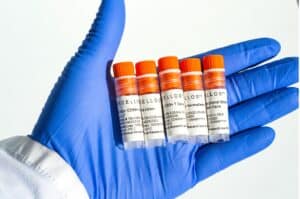A researcher wearing blue nitrile gloves holds five Excellos sample vials with orange caps, containing various isolated immune cells including CD56+ T Cells, CD34+ cells, CD3+ T Cells, and Gamma Delta T cells. Each vial is clearly labeled with product codes and research use designation